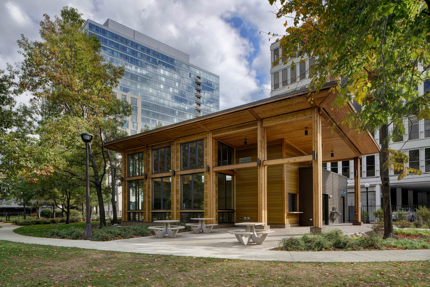 A modern wooden pavilion with large glass windows stands in a park, surrounded by trees and tall office buildings, with picnic tables and a curved walkway in front.