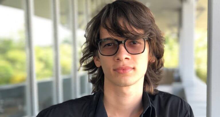 A young person with long brown hair and glasses stands outdoors, wearing a black shirt. Soft natural light and blurred greenery are visible in the background.