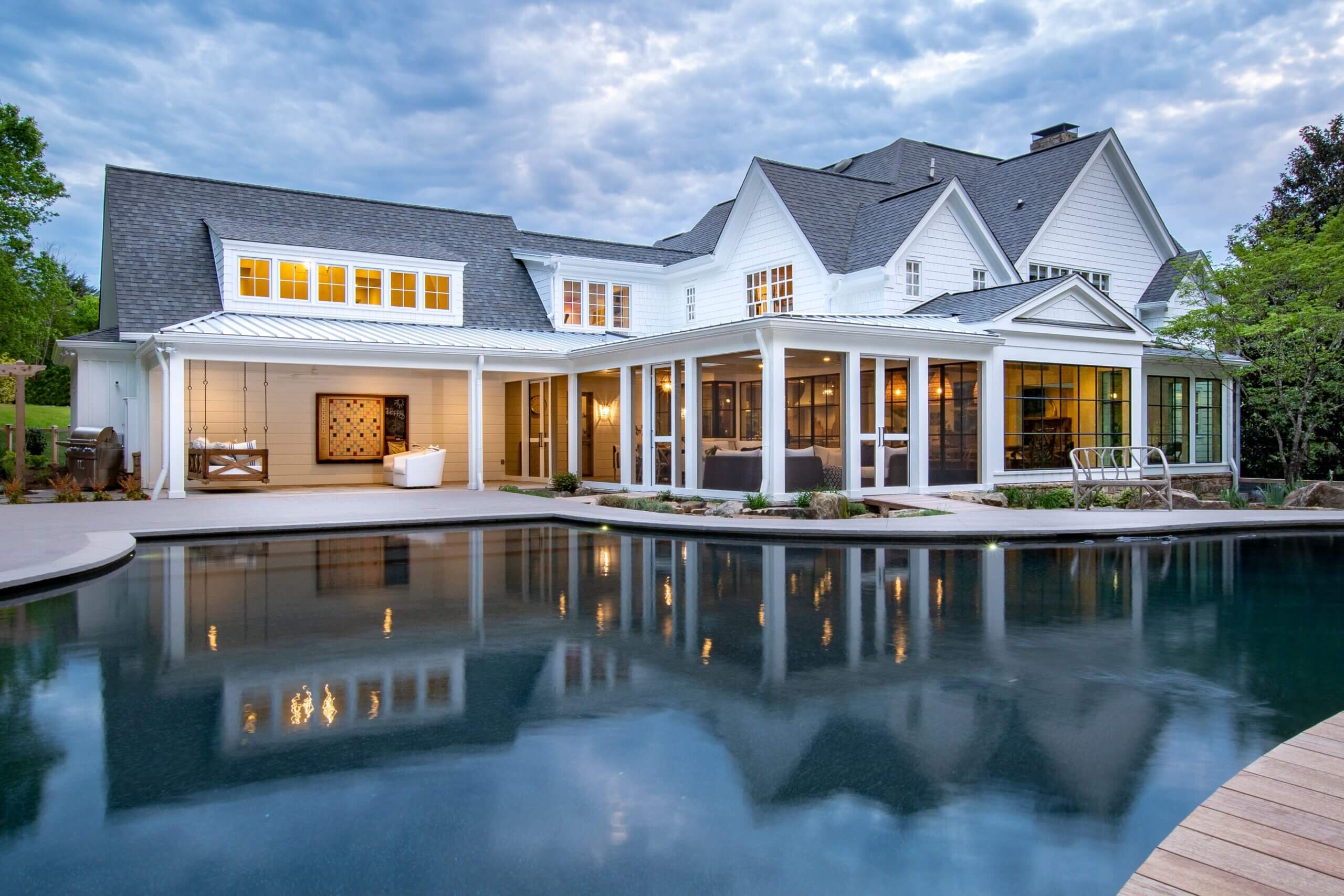 A large, elegant white house with many windows and a spacious covered porch is reflected in a curved swimming pool at dusk, surrounded by landscaped greenery.