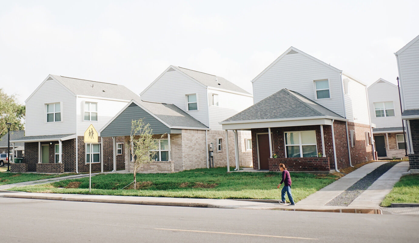 A person walks on a sidewalk past modern, two-story suburban houses with lawns and driveways on a sunny day. A pedestrian crossing sign is visible near the street.