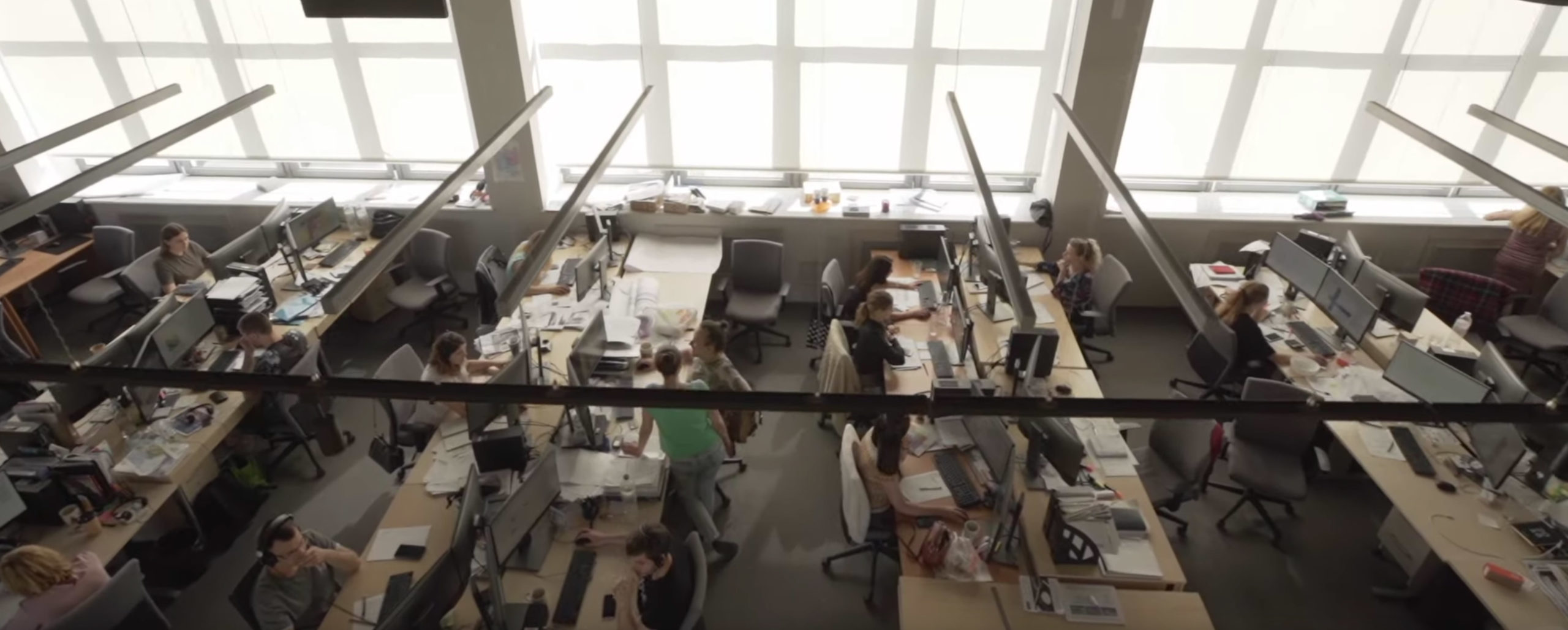 From an aerial view, a bustling office hums with activity, where individuals work at desks adorned with computers and stationery. Large windows let in natural light, highlighting a space enhanced by Archicads design precision and Graphisofts innovative vision. Partitions provide essential privacy.