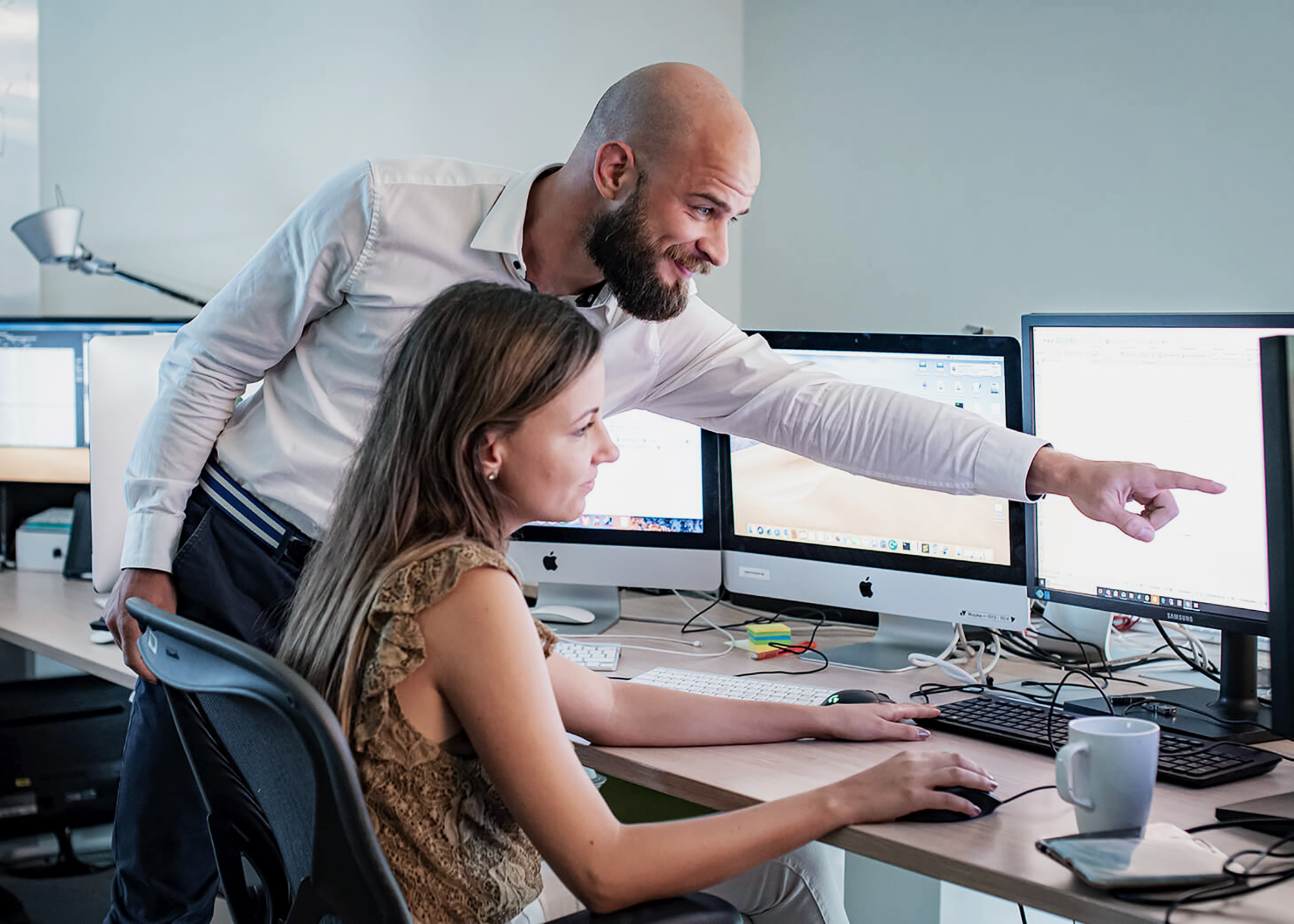 A man and a woman collaborate in an office, immersed in their work with multiple monitors. The man stands, pointing at a screen displaying Archicad, while the woman deftly navigates CAD tools with her keyboard and mouse.