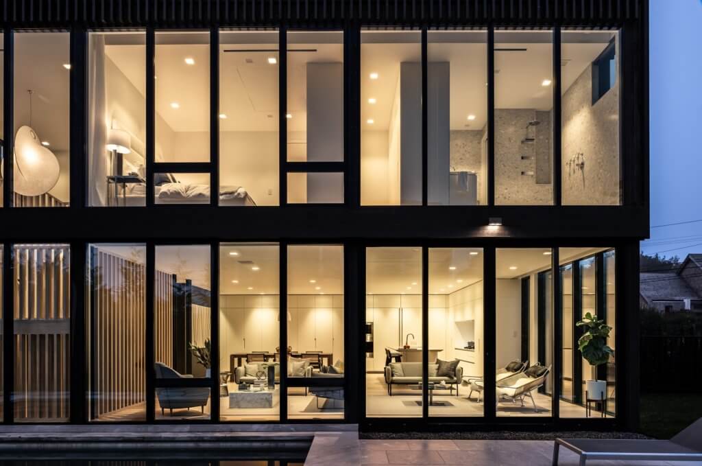 Modern living room with black and white minimalist decor, featuring a black couch, round coffee table, staircase with open risers, wall art, large windows, and a dining area with a plant in the background.