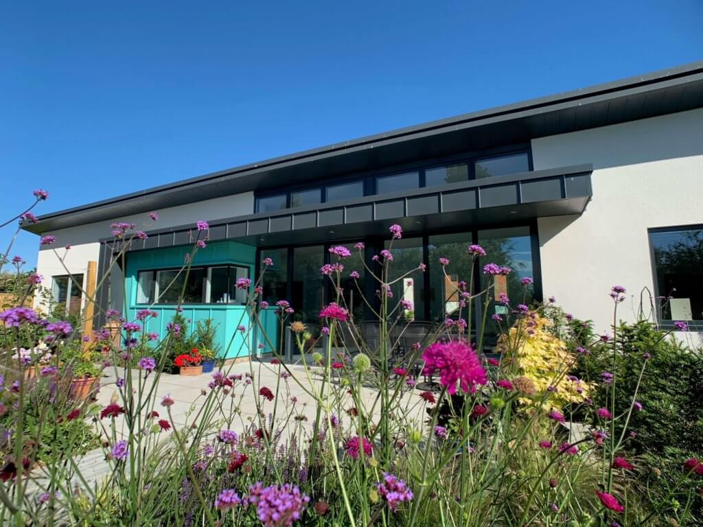 A modern building with a sloped roof and large windows, partially visible behind a garden filled with purple and pink flowers under a clear blue sky, exemplifies an architects vision brought to life through BIM technology.