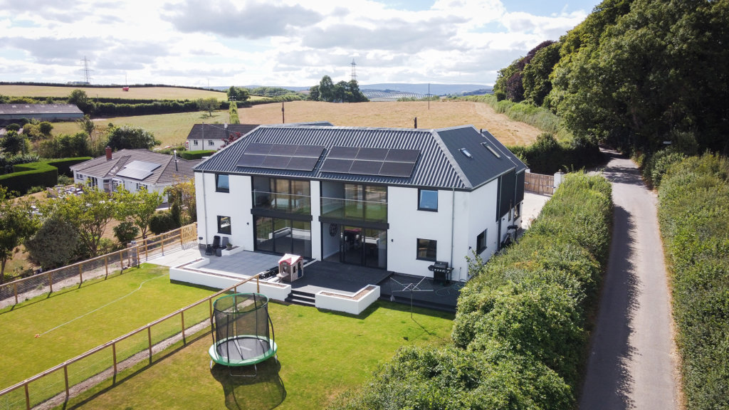 Aerial view of a modern two-story house designed using Archicad, with solar panels on the roof. It features large windows, a lawn with a trampoline, and is surrounded by hedges and trees. A narrow road runs alongside the property, with fields in the background.