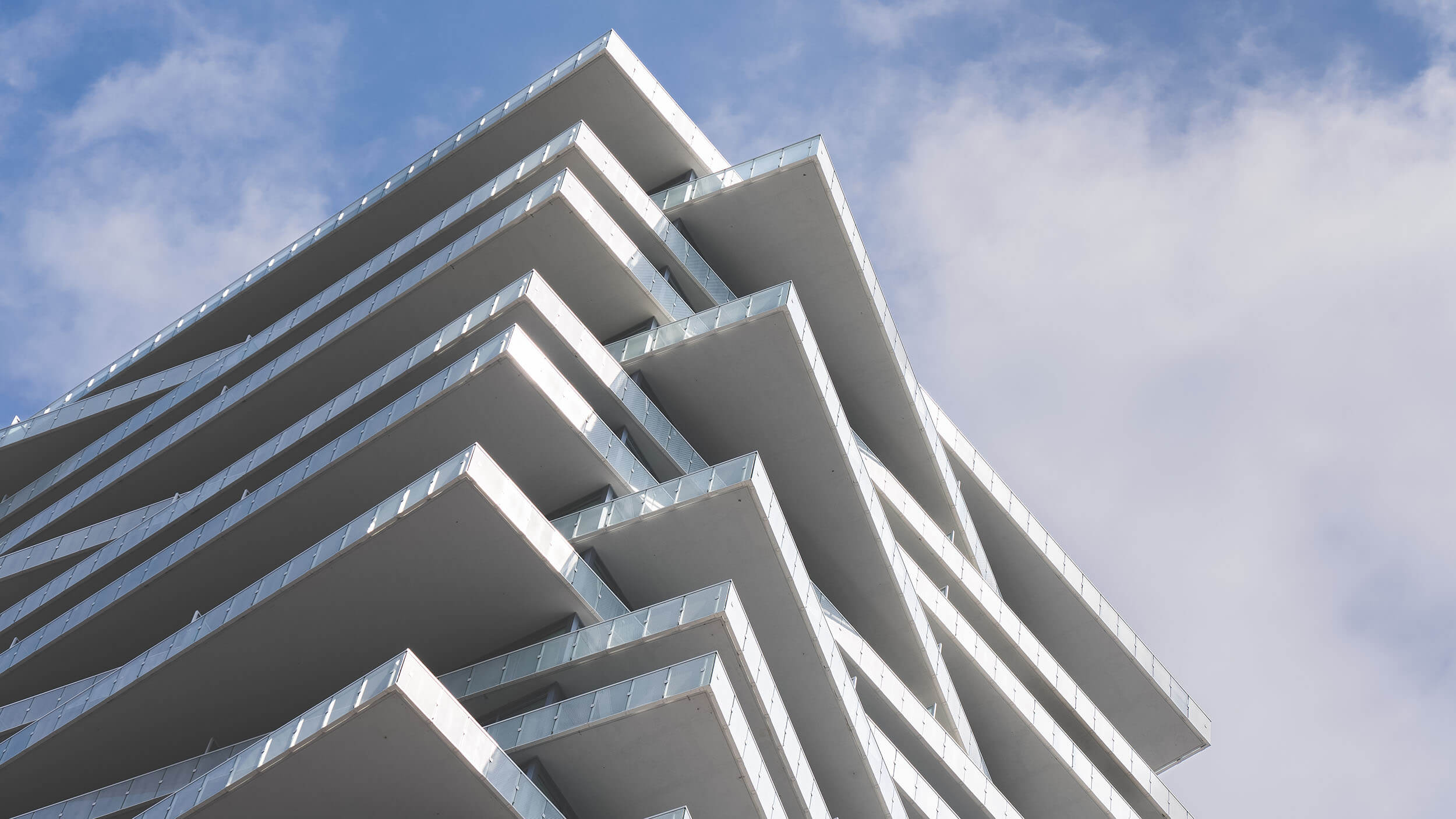 Modern building with staggered, angular balconies against a blue sky with scattered clouds. The white structure creates a geometric, layered appearance.