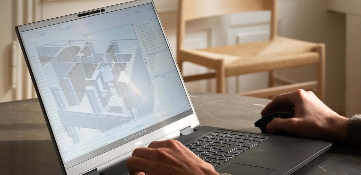 Person using a Dell laptop with architectural design software open, displaying a 3D floor plan. A wooden chair and part of a table are visible in the background.