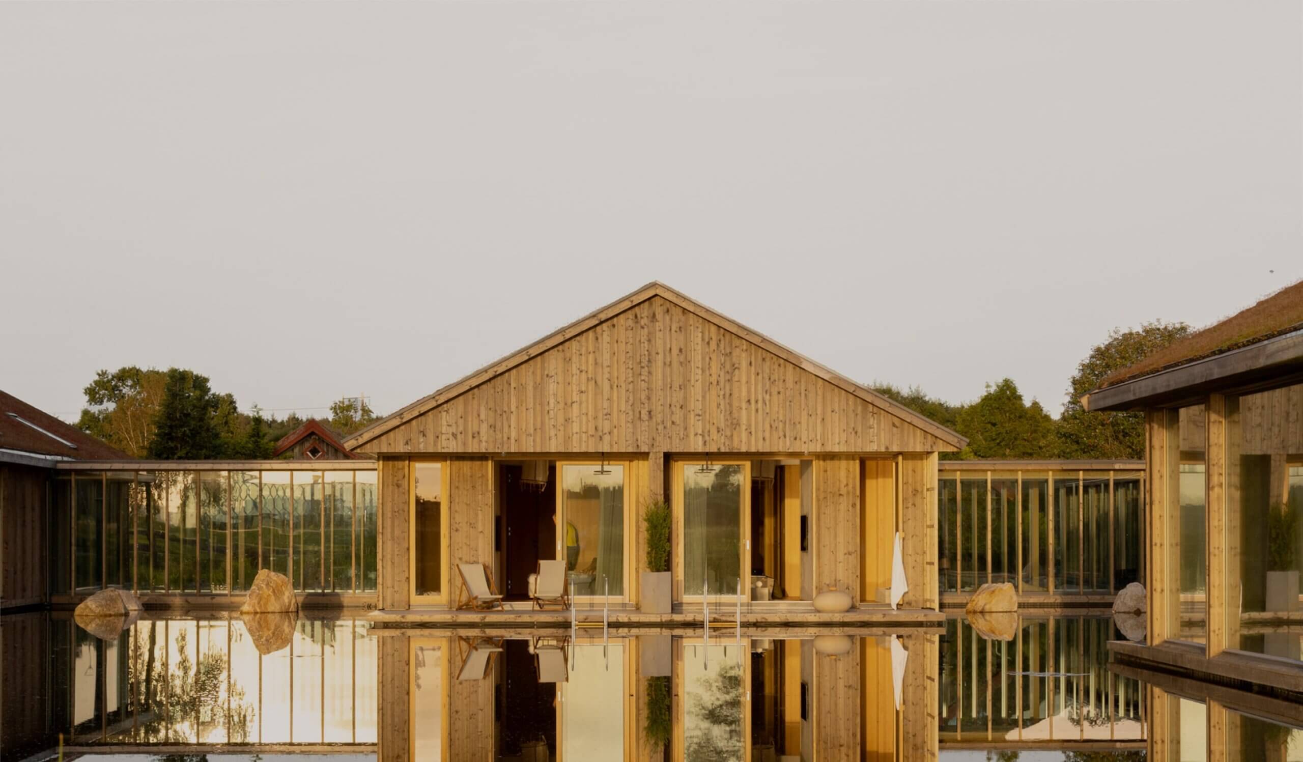 A modern wooden building with large glass windows is reflected in a still pond. The structure has a peaked roof, natural wood texture, and is surrounded by greenery and trees under a clear sky.