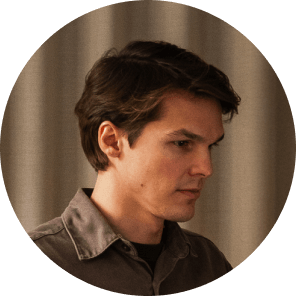 A young man with short brown hair and light skin, wearing a grey collared shirt, is looking down thoughtfully against a neutral, softly lit background.