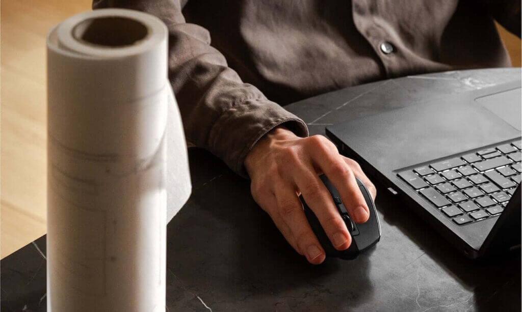 A person wearing a brown shirt uses a black computer mouse next to a laptop on a dark table, with a rolled-up paper or blueprint in the foreground.