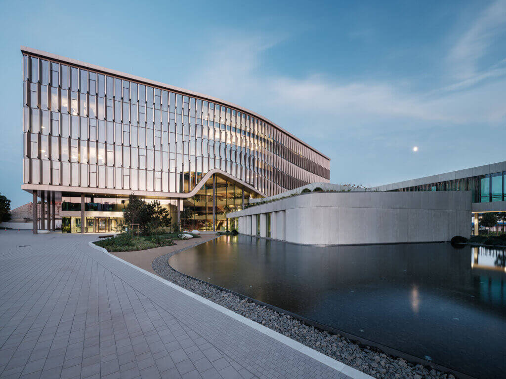Modern glass-fronted building with curved lines, large windows reflecting the sky, and a landscaped area featuring a smooth pond beside a white, angular structure at sunset. The building is Richter Gedeon HQ of Zoboki Design & Architecture and was designed with Archicad using collaborative BIM techniques.
