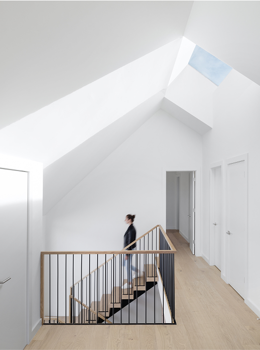 A person ascends a wooden staircase with black railings in a minimalist, white hallway with skylights and light wood floors. Multiple doors line the walls, and natural light brightens the space.