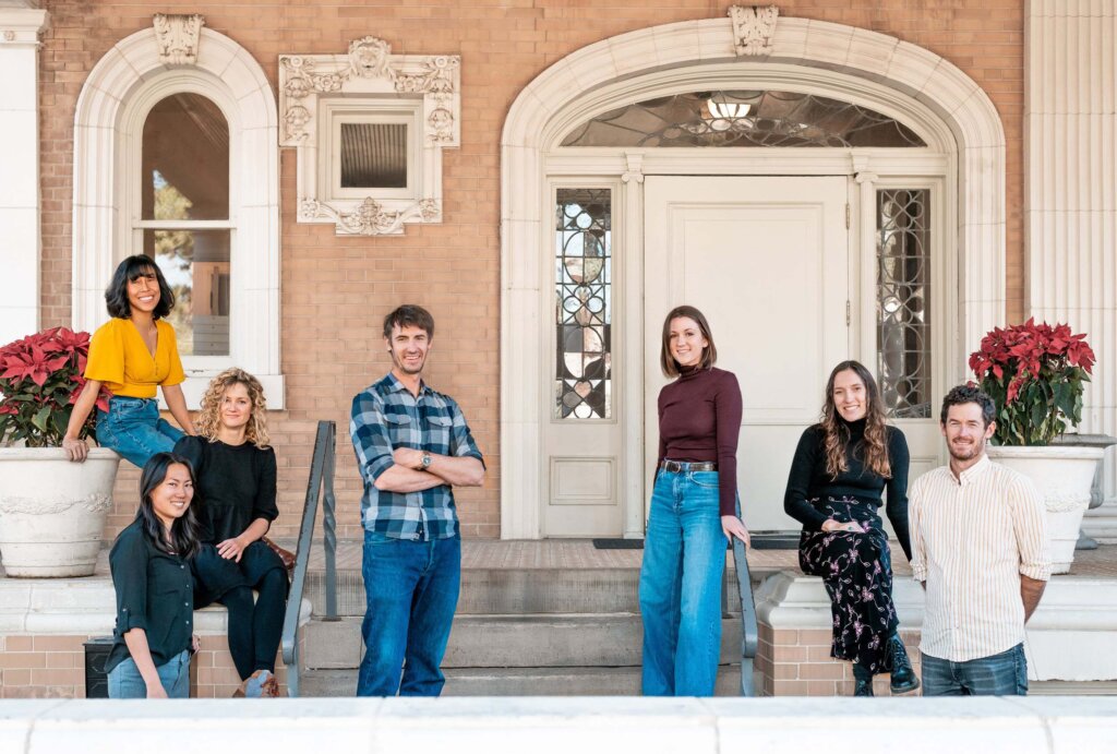 Seven people pose and smile on the steps in front of a building with an ornate entrance and large white door. Two large potted poinsettias sit on either side of the stairs.