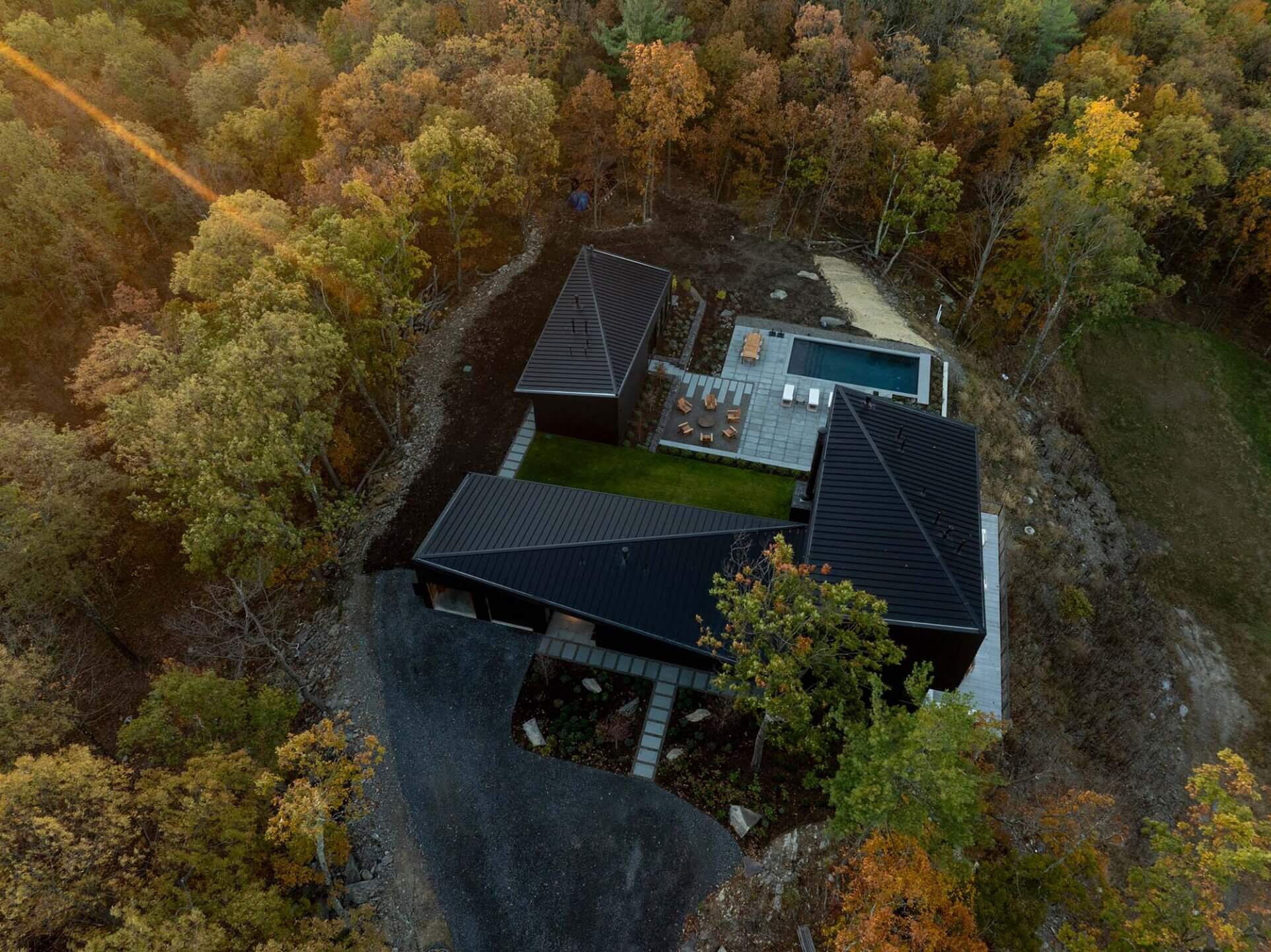 Aerial view of a modern house with a dark roof and central green space, surrounded by autumn trees. The property includes a patio with seating, and a driveway leading to the house through the forested area.