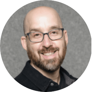 Un hombre sonriente, calvo, con gafas, barba recortada y bigote, vestido con camisa de cuello oscuro, posa delante de un fondo gris liso.