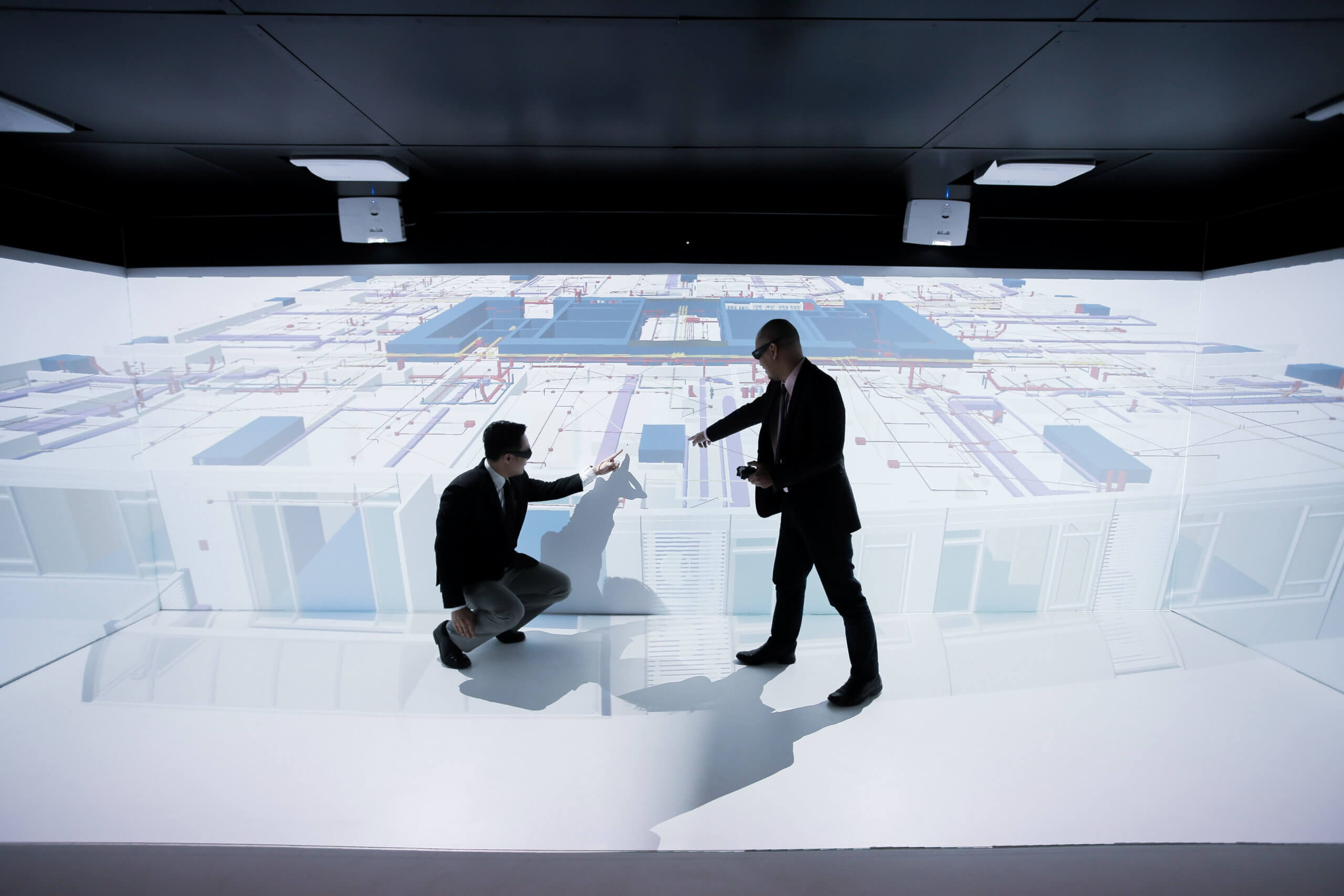 Two people interact in a high-tech room with 3D architectural plans projected on the walls and floor, one person standing and the other crouching as they examine the digital display.