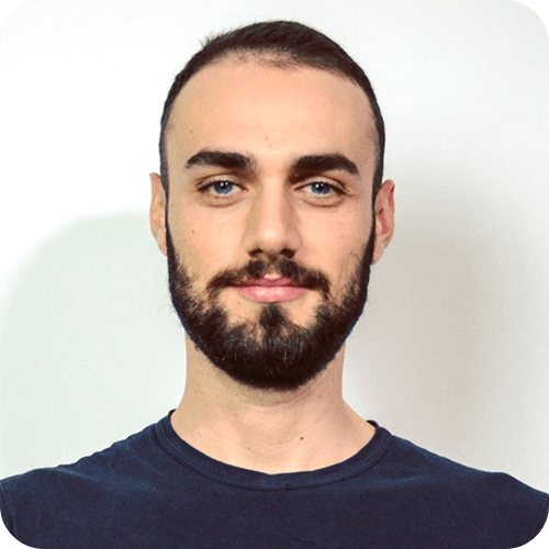A young man with short dark hair and a full beard is facing forward and smiling softly. He is wearing a dark t-shirt and standing against a plain white background.