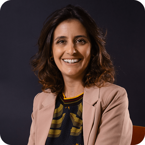 A woman with wavy brown hair smiles at the camera. She is wearing a light pink blazer over a dark shirt with a yellow pattern, and is seated against a dark background.