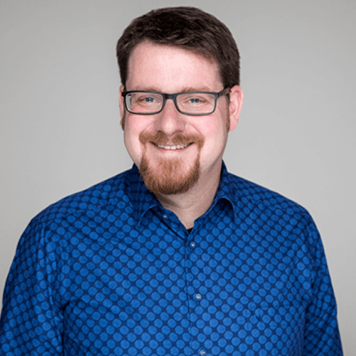A man with short brown hair, a beard, and glasses is smiling. He is wearing a blue shirt with a circular pattern and is standing in front of a plain, light gray background.