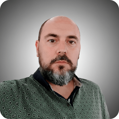 A man with a bald head and a short, graying beard, wearing a green patterned shirt, stands in front of a gray gradient background.