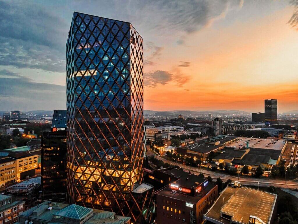 A modern glass skyscraper with a diamond-patterned facade reflects sunset colors, surrounded by city buildings and streets lit by streetlights, under a partly cloudy sky.