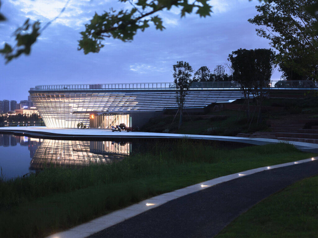 A modern, glass-fronted building with bright interior lights is reflected in a calm lake at dusk. A curved pathway with embedded lights leads through a grassy area toward the building, and trees frame the scene.