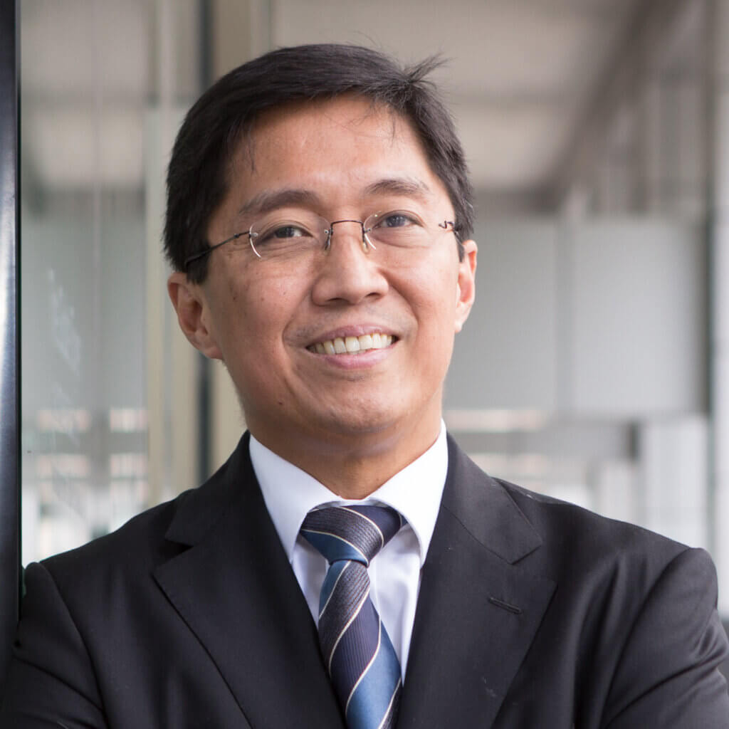 A man in a suit and tie, wearing glasses, stands with arms crossed and smiles confidently in a modern office setting with glass walls.