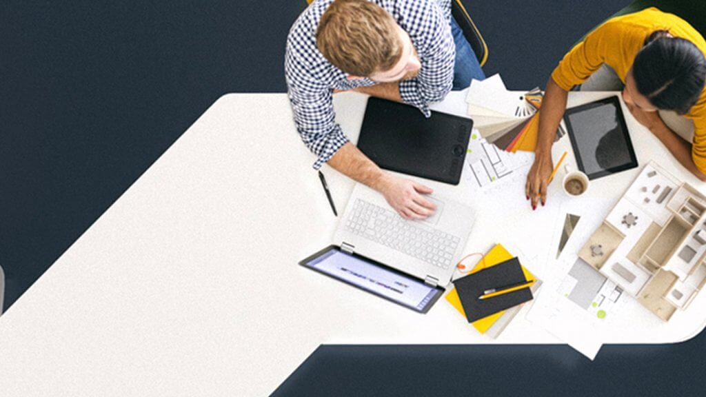 Two people sit across from each other at a white desk, working with a laptop, tablet, papers, a notebook, pencils, and an architectural model, viewed from above.