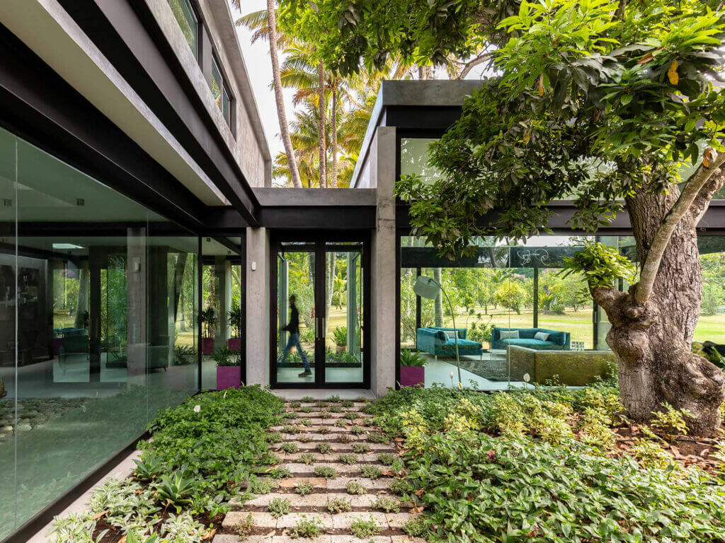 A modern house with large glass windows and doors opens onto a garden with green plants, stone pathways, and a large tree. The interior and outdoor seating area are visible through the glass, surrounded by lush greenery.