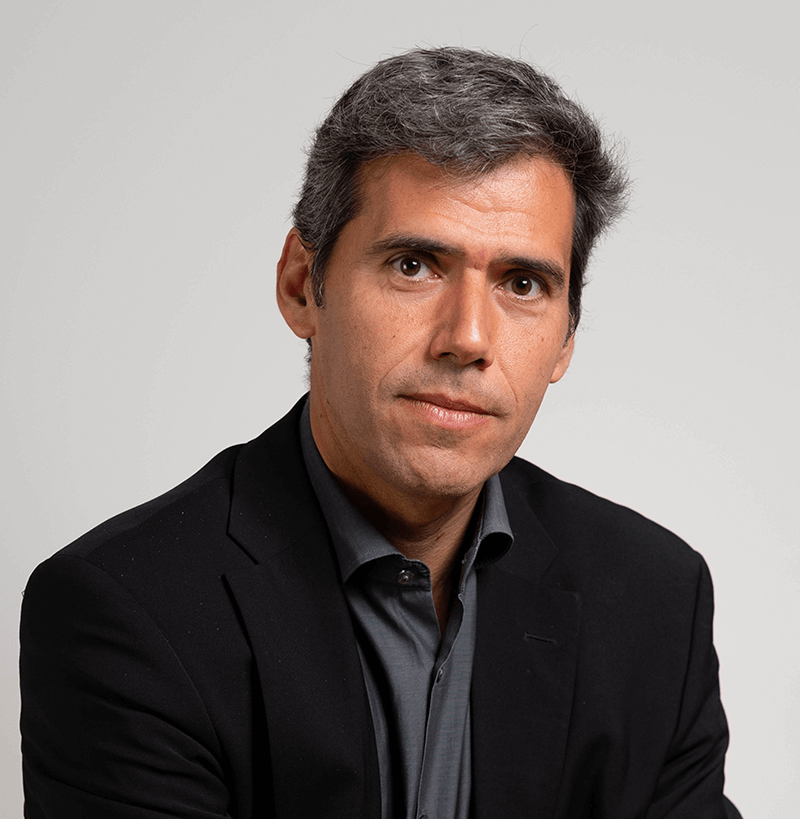 A man with short gray hair wearing a black blazer and dark shirt looks at the camera with a neutral expression against a plain light background.