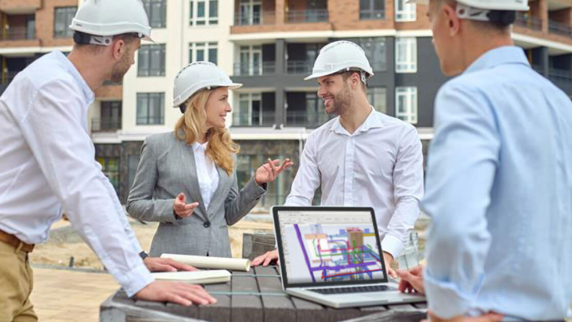 Four people wearing hard hats and business attire stand at a construction site, discussing plans. A laptop displaying a building design is on the table in front of them. Apartment buildings are visible in the background.