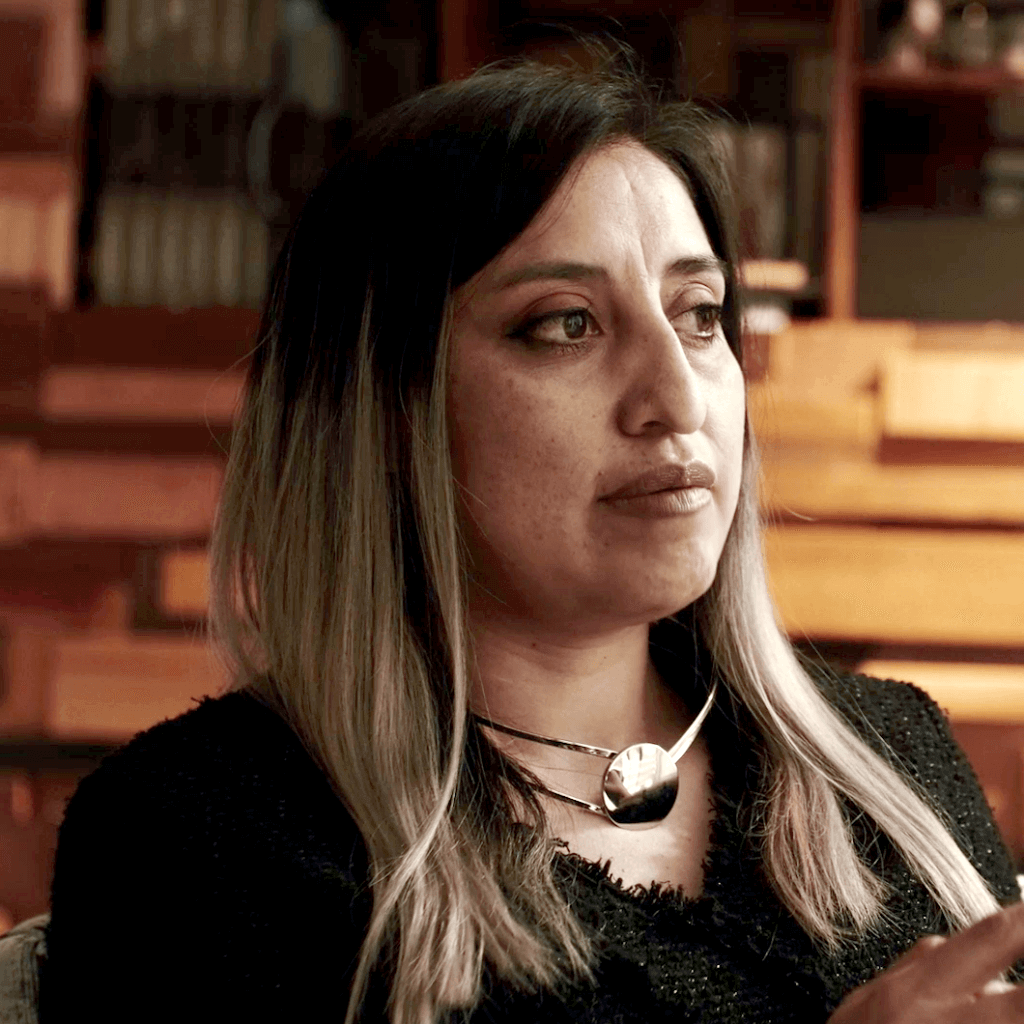 A woman with long, straight, dark-to-blonde ombre hair sits indoors, looking to the side. She wears a black top and a bold, silver necklace. The background shows wooden shelves and warm lighting.