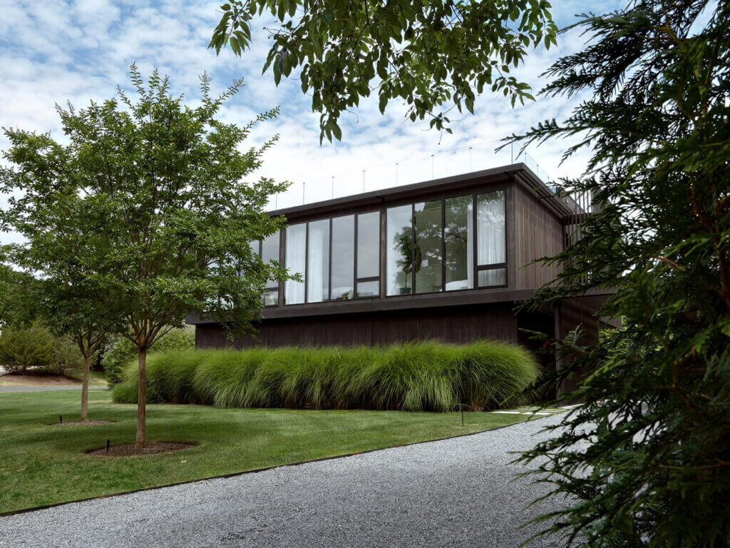 Modern rectangular house with large glass windows, dark wood exterior, and tall grasses surrounding the building. A gravel path curves through a neatly manicured lawn, with leafy trees framing the scene under a partly cloudy sky.