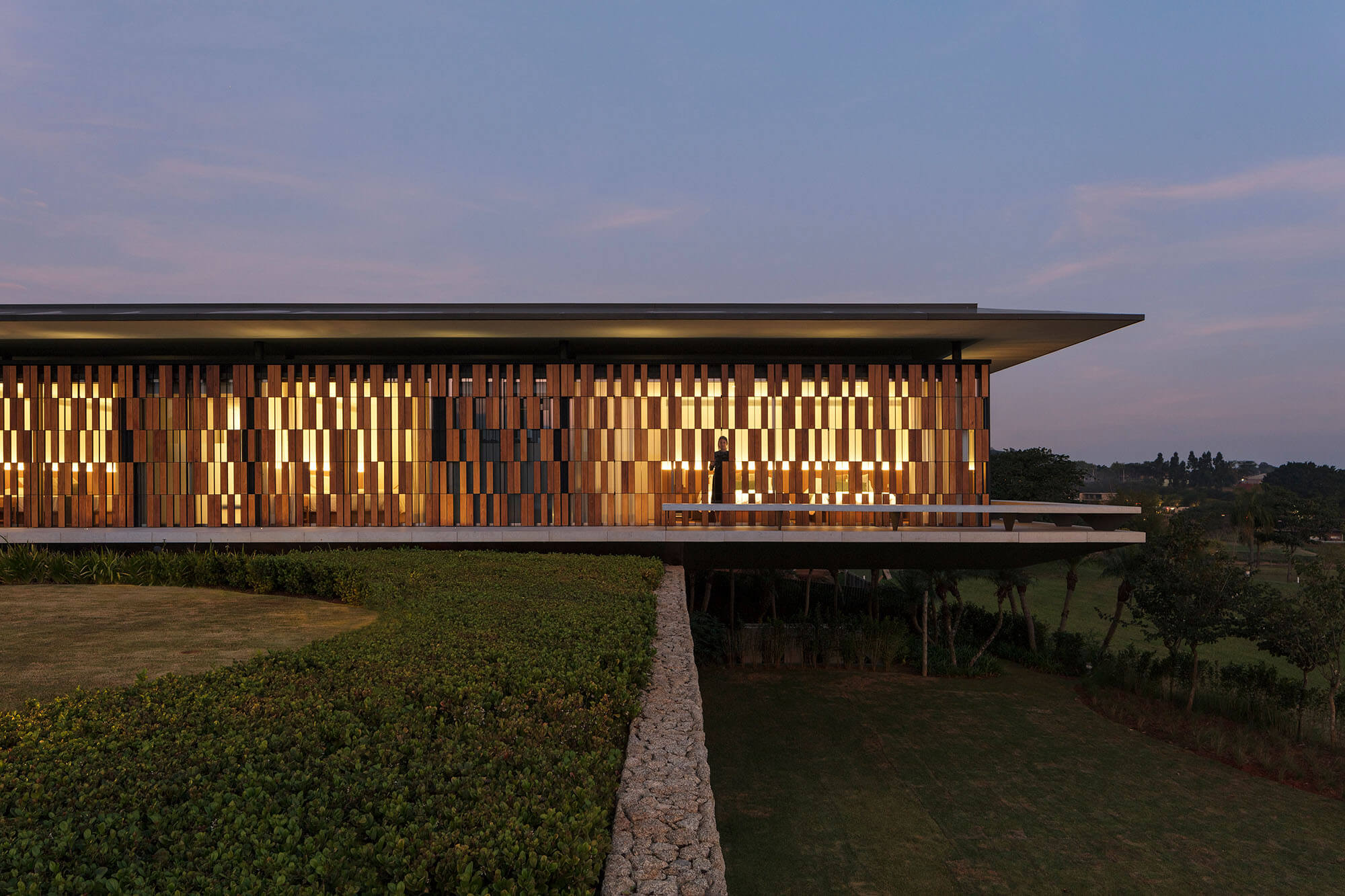 A modern, flat-roofed building with a facade of vertical wooden panels is illuminated at dusk, overlooking a landscaped lawn and greenery, with a stone retaining wall in the foreground.
