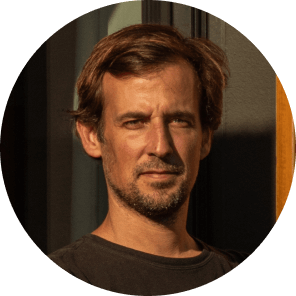 A man with short brown hair and light facial hair stands in sunlight, wearing a dark shirt, looking directly at the camera with a neutral expression. The background is indoors with some shadows.