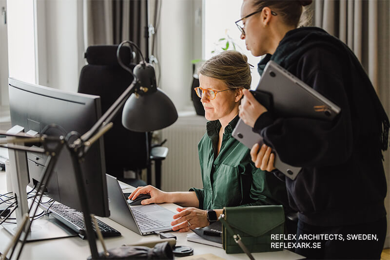 Two women work at a computer in a modern office. One is seated, focused on the screen, while the other stands beside her holding two laptops. Office supplies and natural light are visible. Text reads: Reflex Architects, Sweden.