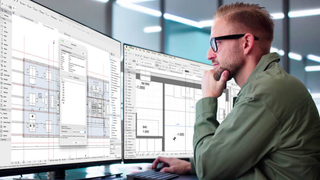 A man wearing glasses and a green shirt works at a desk with two large computer monitors displaying architectural or engineering design software with technical drawings and plans.