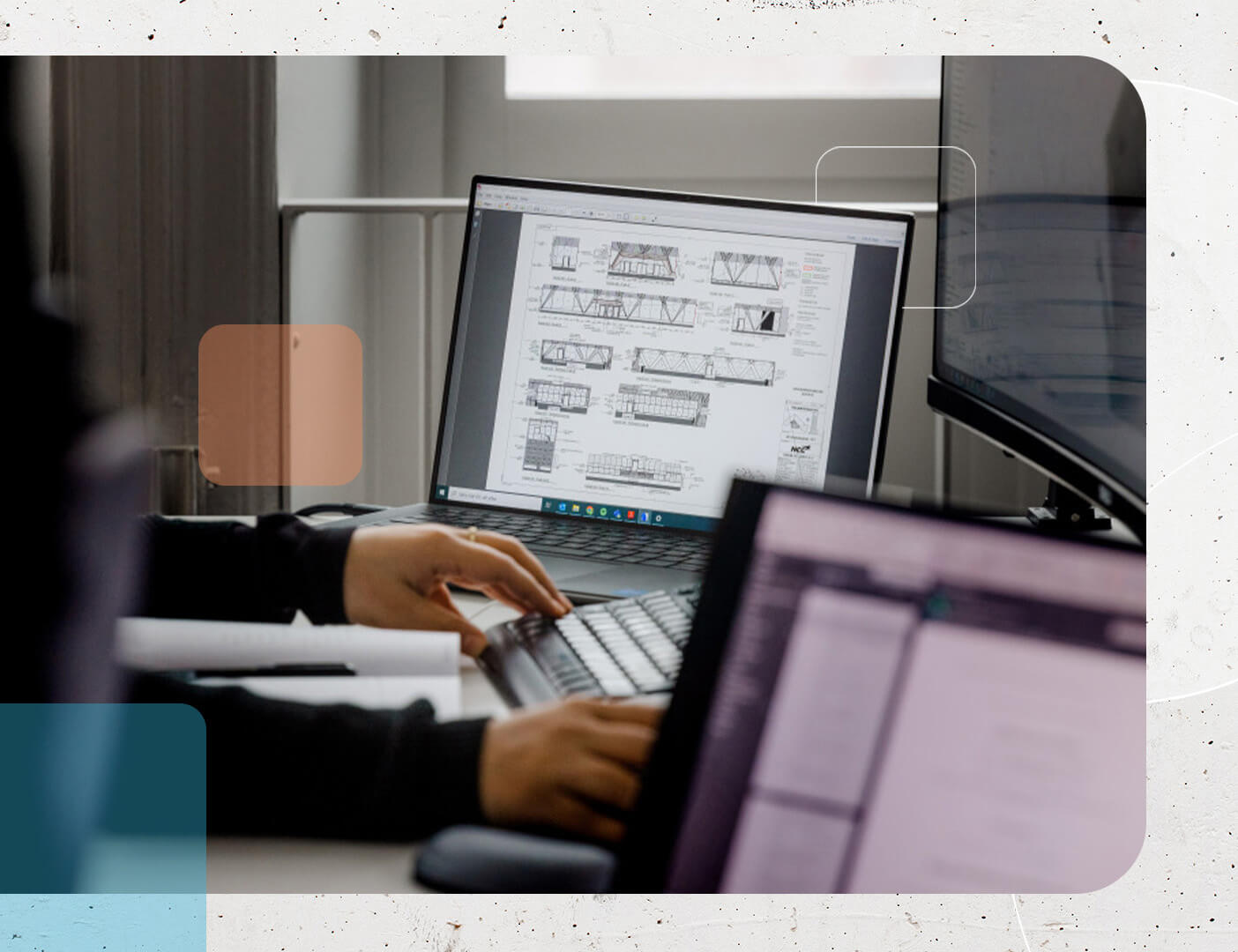 2台のコンピューター・モニターが置かれたデスクで作業する人物。一方のスクリーンには技術的な建築図面が表示され、もう一方にはぼやけたテキストが表示されている。舞台はオフィスかワークスペースのようだ。.