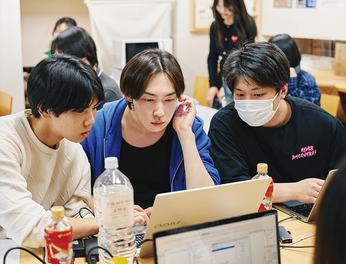 3人の若者が仲良く座り、ノートパソコンの画面に集中している。1人は青いパーカー、もう1人は黒いシャツにフェイスマスク、3人目はクリーム色のセーターを着ている。テーブルの上にはボトルとノートパソコンが置かれ、背後では他の人々が作業をしている。.