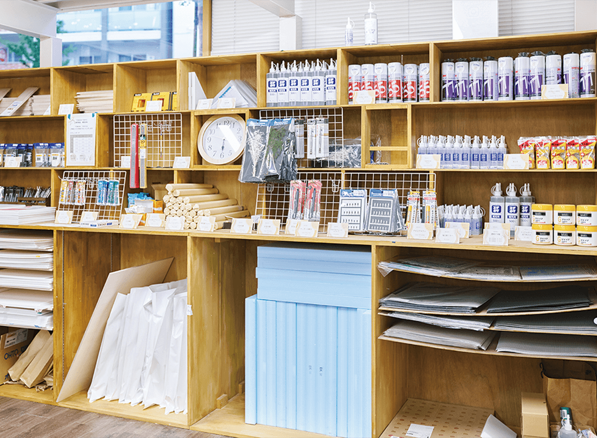 店内の木製の棚には、のり、絵の具、定規、ロール紙、時計など、さまざまな文房具や画材が整然と並べられている。.