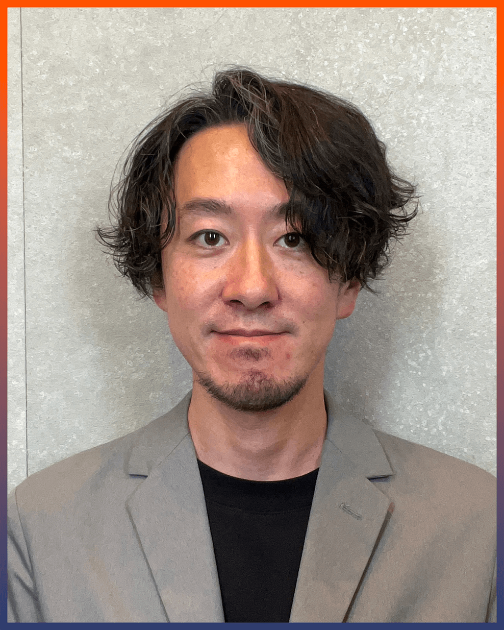 ウェーブのかかったミディアムヘアの黒髪で、明るい顔立ちの男性が、黒いシャツの上にグレーのブレザーを着て、ライトグレーの質感の背景の前に立っている。.