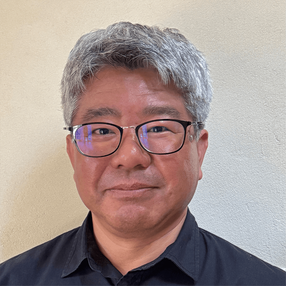 眼鏡をかけ、濃い色の襟付きシャツを着た灰色の短髪の中年男が、淡い色の無地の壁の前に立って微笑んでいる。.