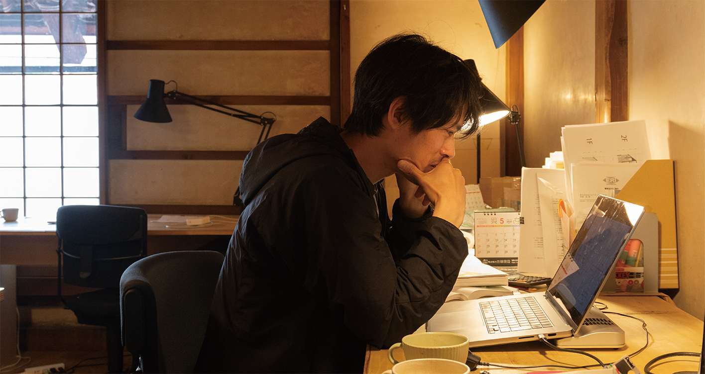 薄暗い部屋のデスクに座り、ノートパソコンに集中している人。ワークスペースには書類、コップ2つ、カレンダーが置かれ、デスクランプの暖かい光が居心地の良い雰囲気を醸し出している。.