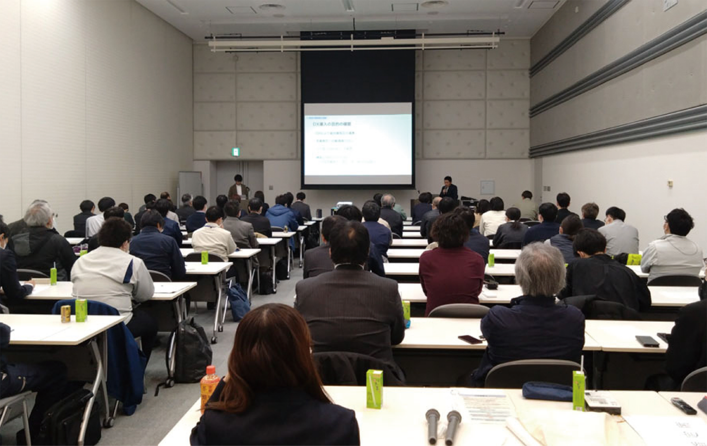 会議室のテーブルに座った一団が、プレゼンテーションが映し出されたスクリーンに向かっている。ほとんどの出席者は前を向いており、部屋は中間色の壁で明るい。.