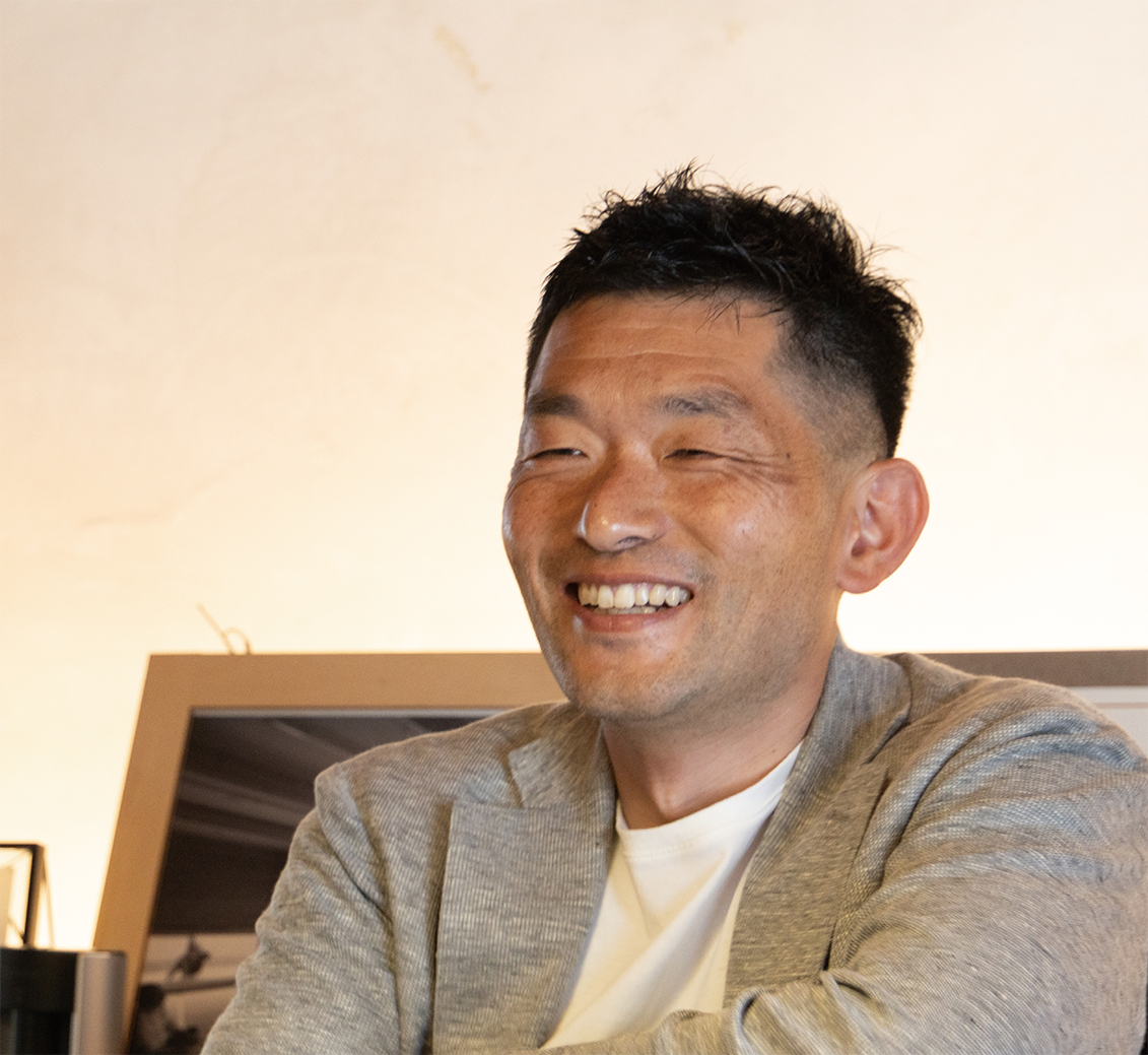 黒髪のショートヘアにライトグレーのブレザーを羽織り、白いシャツを着た男性が温かな笑みを浮かべている。額縁に入った写真とニュートラルな壁を背景に、室内で座っている。.