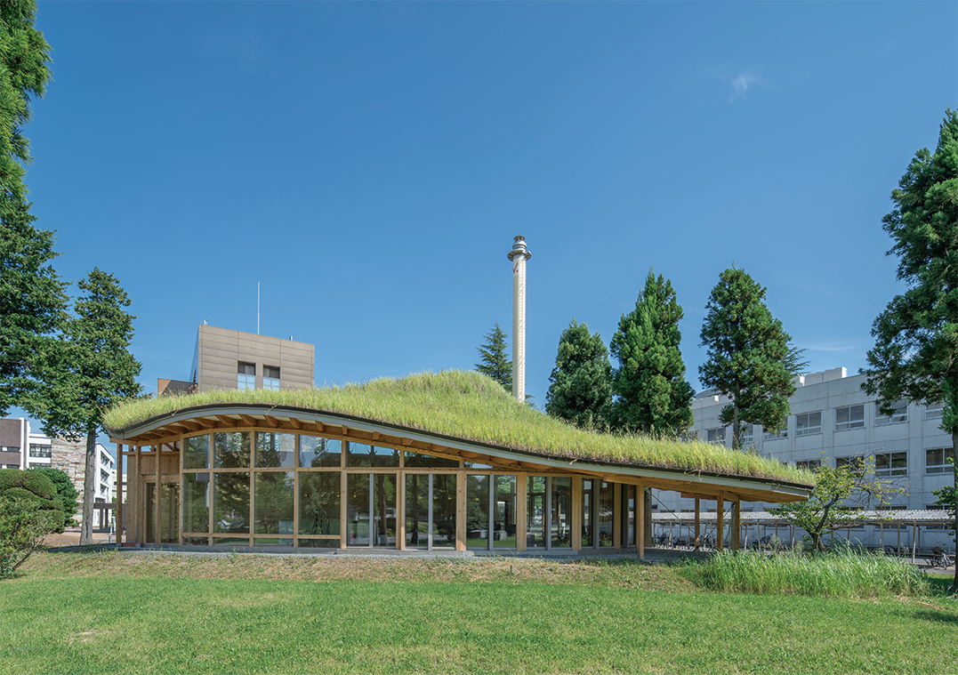 大きなガラス窓と芝生に覆われた波打つ緑の屋根を持つモダンな建物が、澄み切った青空の下、木々と芝生に囲まれている。.