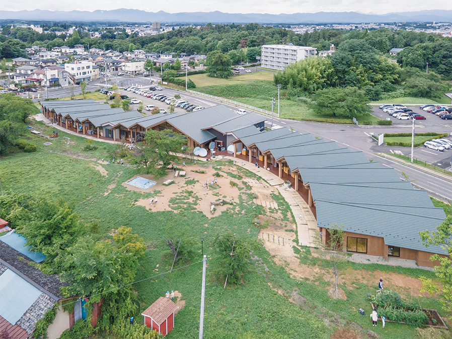 緑豊かな半農村地帯に建つ、ジグザグ型の屋根を持つモダンな平屋建ての建物の航空写真。周囲には樹木や空き地があり、近隣の住宅地や駐車場もある。.