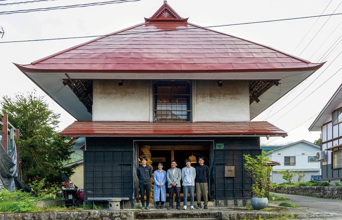 大きな赤い屋根を持つ伝統的な和風建築の前に6人が立っている。建物の下半分は黒っぽい木の壁で、上半分は白い壁。舞台は住宅街のようだ。.