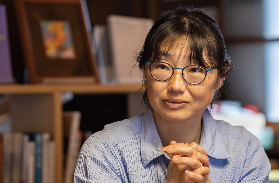 眼鏡をかけ、青いシャツを着た女性が本棚の前に座り、両手を合わせて物思いにふけっている。背景は本や額に入った絵など、柔らかくぼかされている。.
