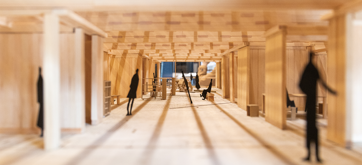 木で作られた建築模型のクローズアップ写真。円柱のあるホールと、空間全体に配置された小さな黒い人型。.